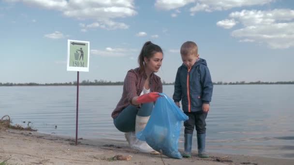 Miljö volontärer, är ung kvinna överväga med unge pojke hur mycket skräp samlas in i sopsäck medan rengöring stranden från plast skräp och ge tummen upp nära floden — Stockvideo