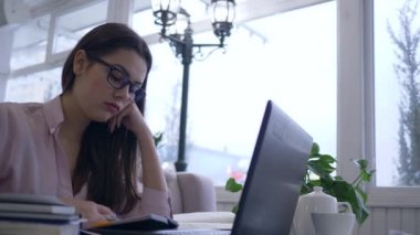 Yorgun, çok çalışan bir bayan öğrenci gözlük takıp uzaktan eğitim sırasında online ders alarak dizüstü bilgisayarla ders çalışıyor ve notlar alıyor.