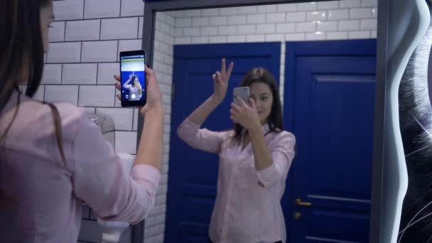 Aantrekkelijk meisje poseren in de voorkant van spiegel voor selfie foto op mobiele telefoon in toilet — Stockvideo