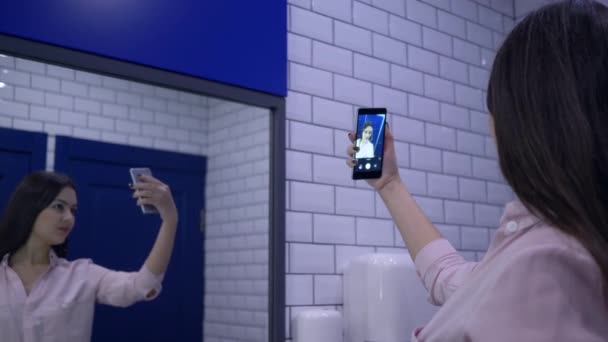 Hermosas mujeres es fotografiado en el teléfono inteligente de pie delante del espejo en el baño — Vídeo de stock