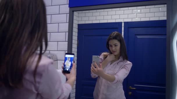 Blogging, young woman records video on mobile phone standing in front of mirror in lavatory — ストック動画