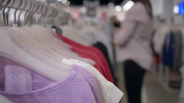 Clothes shop, fashionable new clothing hanging on hangers and shopper woman in unfocused background chooses purchases in store — Stock Video