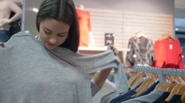 Mutlu gülümseyen alışveriş delisi kız yeni şık kıyafetler seçiyor ve indirimler sırasında moda mağazasında aynaya bakıyor.