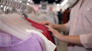 Alışverişçi kadın, indirimler sırasında moda mağazasında askılarda yeni şık kıyafetler seçiyor, elleri odaklanmamış arka planda.