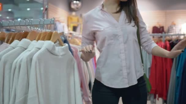 Магазины продаж, привлекательная женщина выбирает и пробует новую одежду перед зеркалом в магазине моды во время скидок в торговом центре — стоковое видео