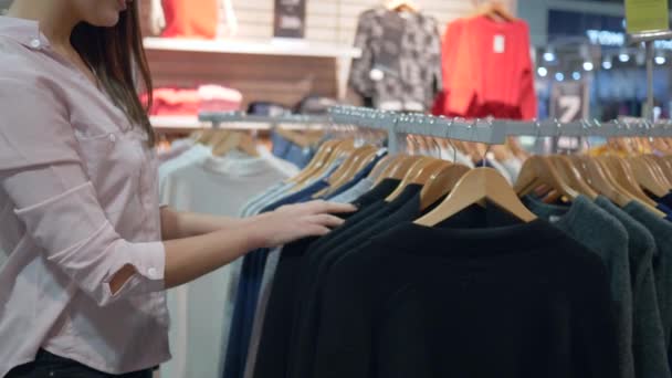 Sonriente shopaholic chica elige nueva ropa en perchas y probándose a sí mismo en la tienda de moda durante los descuentos en el centro comercial — Vídeos de Stock