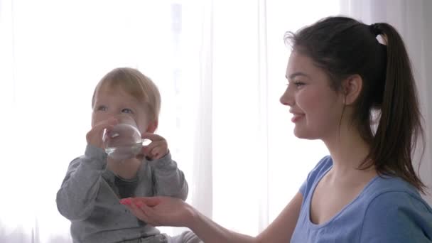 Primer niño éxitos, niño feliz bebe independientemente agua mineral de vidrio al lado de la joven madre sonriente en luz natural — Vídeo de stock