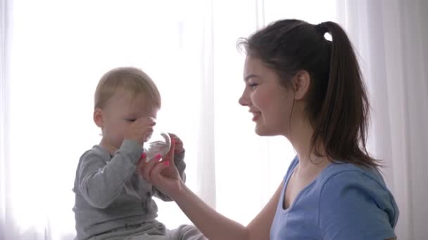 Cuidado materno, feliz garotinho rindo bebendo água mineral pura de vidro de mãos de mãe sorridentes e se alegra em saciar a sede na luz natural no quarto — Vídeo de Stock