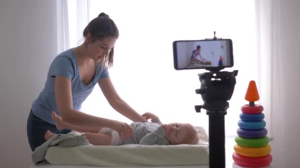 Cuidado del bebé, feliz blogger mamá cambia la ropa del niño mientras graba video de entrenamiento en el teléfono móvil para los suscriptores en las redes sociales en streaming en vivo — Vídeo de stock