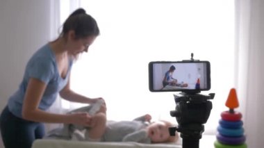 İş blogcusu, genç anne vlogger, canlı yayında sosyal ağlardaki takipçiler için akıllı telefondan eğitim videosu kaydederken yeni yürümeye başlayan çocuğun kıyafetlerini değiştiriyor.