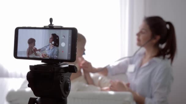 Entrenamiento en línea, médico de familia mujer con estetoscopio escucha los latidos del corazón y el aliento del niño pequeño paciente durante la grabación de la lección de vídeo en directo transmitido en el teléfono móvil para los seguidores — Vídeo de stock