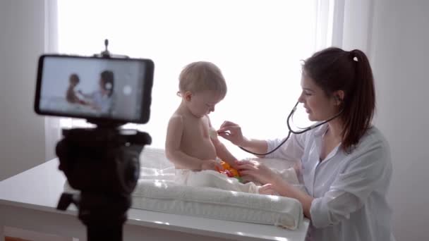 Lección de video, blogger madre médico examinando hijo usando estetoscopio y conduce transmisión de aprendizaje en vivo en el teléfono inteligente para los seguidores en las redes sociales — Vídeo de stock