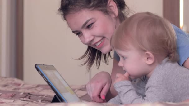 Modern çocuk gelişimi, mutlu annesi olan sevimli bir çocuk. Dijital tabletle yatağında yatıyor. — Stok video