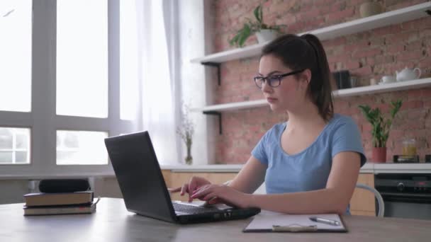 Vzdělávání, studentka v brýlích používat počítač notebook ráno a pije kávu s bílým šálkem pak píše poznámky ve schránce sedí u stolu doma — Stock video
