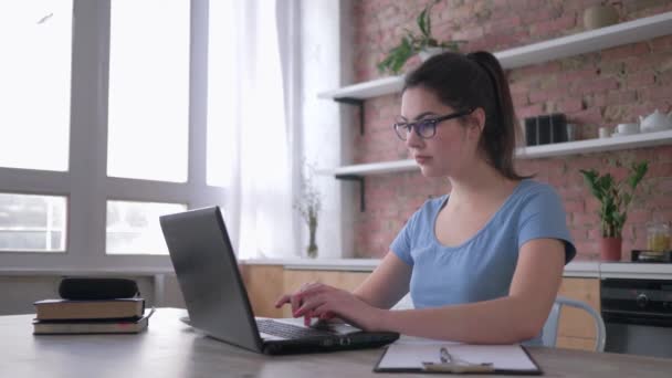 Online training, knappe jonge vrouw in bril gebruik laptop en schrijft notities in klembord zitten aan het bureau in de keuken — Stockvideo