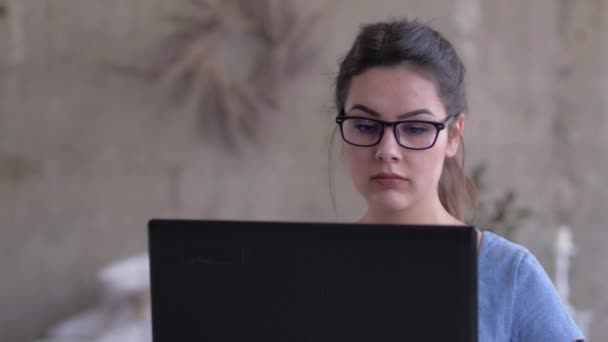 Портрет девушки-фрилансера в очках, работающей на ноутбуке онлайн, сидящей дома крупным планом — стоковое видео