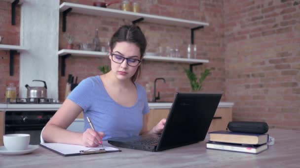 Remote job, jonge vrouw in bril schrijft notities in klembord en typen op laptop toetsenbord in slow motion zitten aan tafel in de keuken — Stockvideo