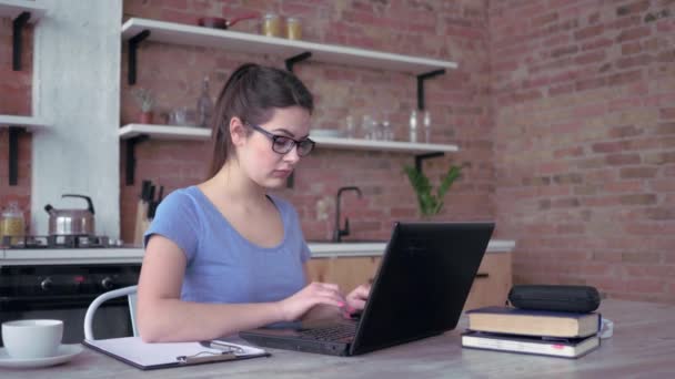 Genç modern kadın evde çalışıyor, gözlüklü kız dizüstü bilgisayarda klavyeyle yazı yazıyor ve mutfaktaki masada oturan panoya notlar yazıyor. — Stok video