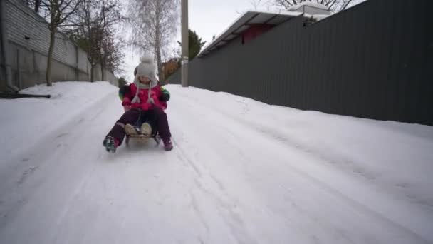 Зимние детские развлечения, счастливый мальчик и девочка катаются на санях по заснеженной дороге — стоковое видео