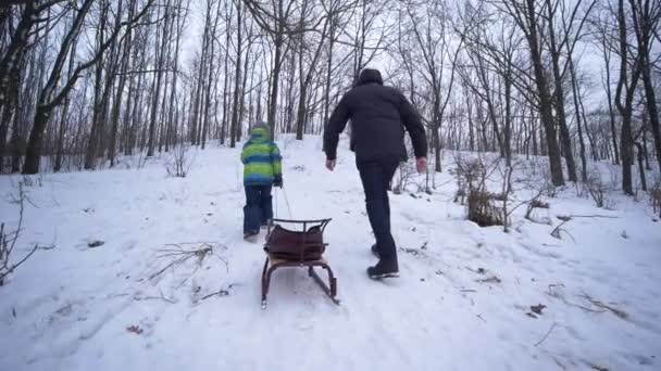 Материнська підтримка, батько допомагає синові підніматися на засніжену гору зі сідлами в руках взимку — стокове відео