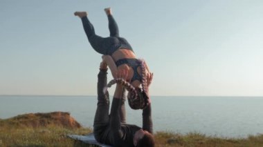 Açık havada akro yoga, takım elbiseli kız deniz ve gökyüzünün arka planındaki çayırda erkek arkadaşının elleri ve bacakları üzerinde dengede duruyor.