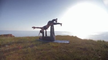 Atletik çift dağda akro yoga yapıyor, dişi akrobat erkek ortağının elleri ve bacakları üzerinde dengede duruyor gökyüzü ve denizin arka planında güneş ışığı altında.