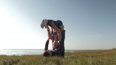 Akroyoga yapan atletik kadın, erkek arkadaşının ellerinde gökyüzünün arka planında çayırda denge kuruyor.