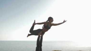 Çayırda akro yoga yapan çift, spor takım elbiseli genç kadın deniz ve gökyüzünün arka planında erkek ortağının bacaklarında dengede duruyor.
