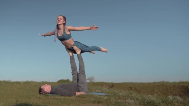 Пара делает акробатические упражнения на лугу, молодая белая женщина в спортивном костюме балансирует на ногах своего партнера-мужчины на фоне неба — стоковое видео