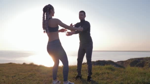 Здоровый образ жизни, пара атлетичных женщин и мужчин, держащихся за руки и одновременно приседающих на солнце на природе — стоковое видео