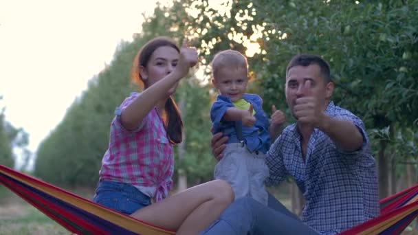 Портрет счастливой сельской семьи, улыбающиеся мама и папа с маленьким сыном давая большой палец вверх во время отдыха в гамаке — стоковое видео