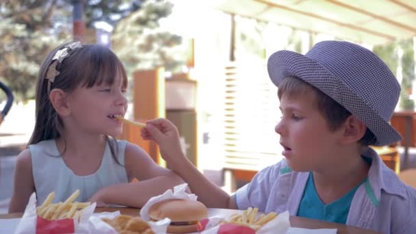 Нездоровое питание для детей, заботливый милый мальчик кормит подружку картошкой фри во время обеда в кафе быстрого питания на улице — стоковое видео
