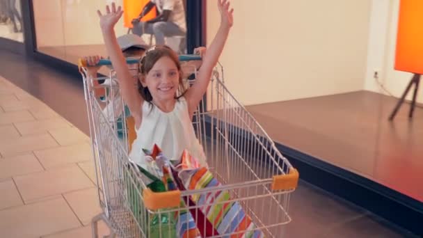 Stagione di sconti, ridere i bambini si divertono nei carrelli dello shopping nel centro commerciale e passare davanti alle vetrine delle boutique — Video Stock