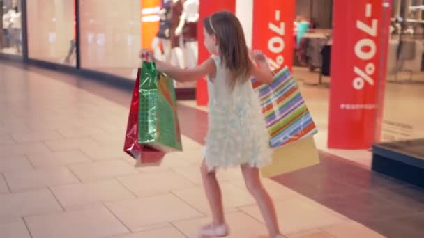 Jour de shopping, fille heureuse enfant avec des paquets dans les mains traverse le centre commercial après l'achat dans les boutiques de mode — Video