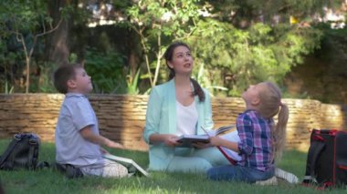 İlkokul, mutlu liseli çocuklar ve özel öğretmen kadınla birlikte olan kız ve erkek öğrenciler çim üzerinde eğitim sırasında kitap okuyup sohbet ediyorlar.
