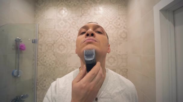 Shaving, attractive male with Electric Razor shaves stubble on face in front of mirror at washroom — Stock Video