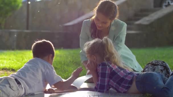 Desarrollo infantil, mujer joven leer libro interesante para los niños sentados en el césped verde en la naturaleza en la luz soleada — Vídeo de stock