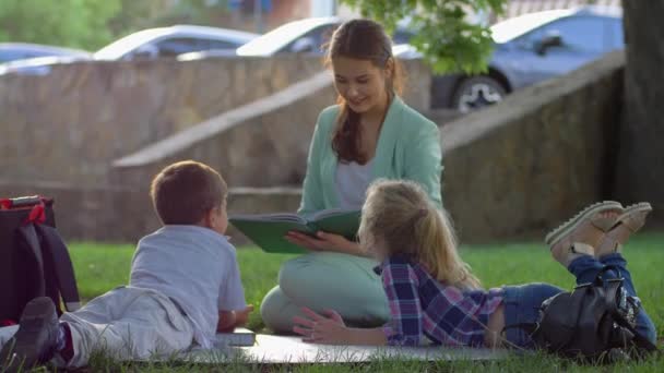 Buitenschoolse activiteiten, schattige kinderen luisteren naar vrouw lezen boek na school zitten op groen gras in de natuur in zonnig licht na schoollessen — Stockvideo