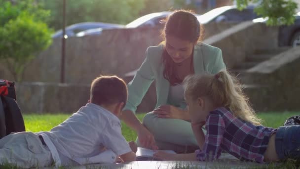 Gelukkige jeugd, lachende moeder lees boek voor kleine jongen en meisje zitten op groen gazon in de natuur in zonnig licht — Stockvideo