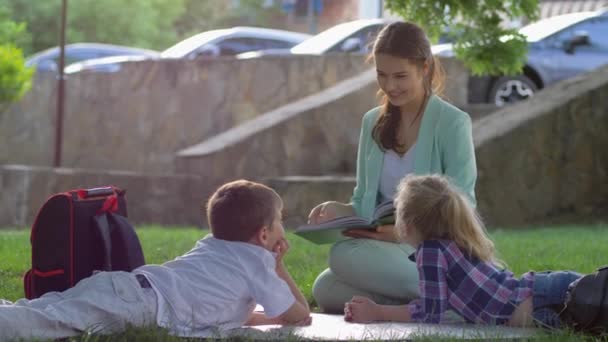 Маленькие школьники слушают женскую книгу для чтения, сидя на зеленой лужайке на природе в солнечном свете после окончания школы — стоковое видео
