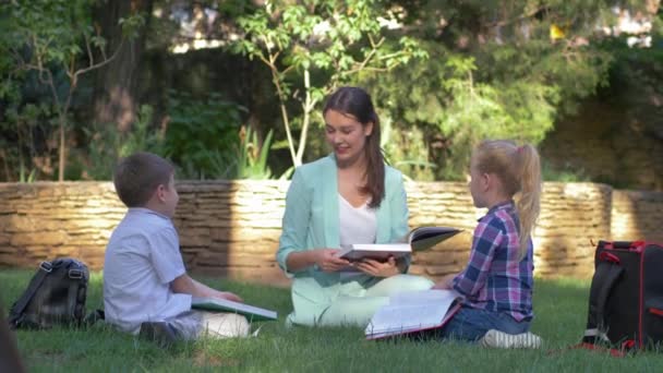 Métodos de educação modernos, alunos colegas menino e menina com tutor feminino ler livro escolar e bate-papo durante a lição sobre gramado — Vídeo de Stock
