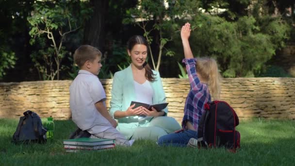 Молода вчителька проводить урок на відкритому повітрі для маленького хлопчика і дівчинки з книгами в руках, сидячи на траві — стокове відео