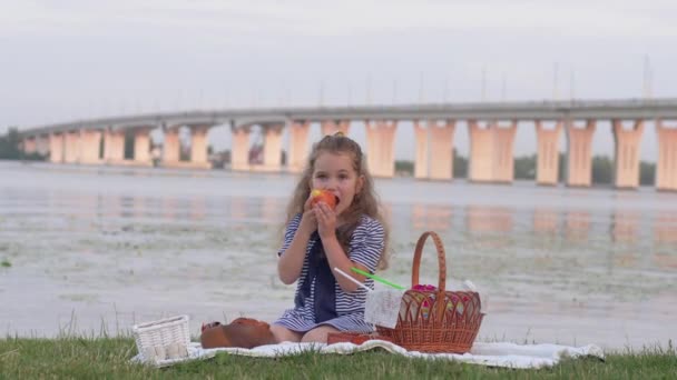 Little cute child girl eating fresh fruit during picnic near river on background of bridge — Stock Video