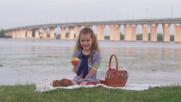 Liten leende flicka äter färskt äpple och ger tummen upp under picknick i naturen nära floden — Stockvideo