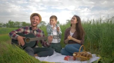 Neşeli baba müzik aleti çalarken anne ve oğlu yeşil alanda aile pikniğinde eğleniyor ve alkışlıyorlar.