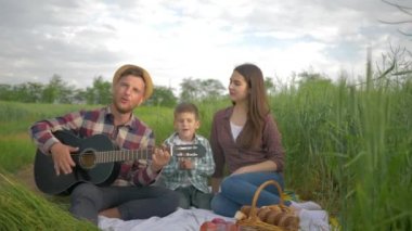 Eğlenceli aile tatili, neşeli erkek müzik aleti çalarken çocuklu kadın yeşil alanda piknik yaparken şarkı söyleyip alkışlıyor.