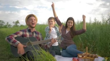 Gülümseyen neşeli baba gitar çalarken, anne ve çocuk yeşil alanda aile pikniğinde dans edip alkışlıyorlar.