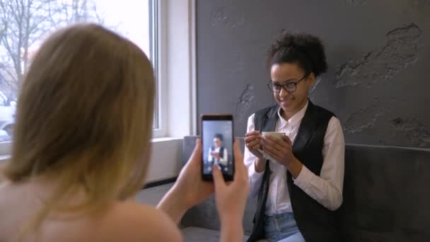 幸せな混合人種の友人、若いアフリカ系アメリカ人の女性はガールフレンドとカフェでリラックスしながらスマートフォンで撮影されます — ストック動画