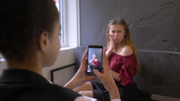 Amigos multi-étnicos felizes, jovem mulher de pele escura fotografias namorada com xícara de chá na mão no telefone móvel no restaurante — Vídeo de Stock