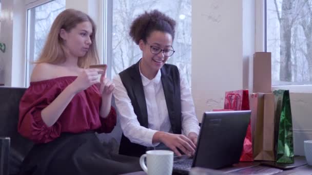 Інтернет-покупки, усміхнені багатоетнічні друзі дівчата використовують пластикову картку і ноутбук для оплати онлайн-покупки, сидячи в кафе з великою кількістю пакетів — стокове відео